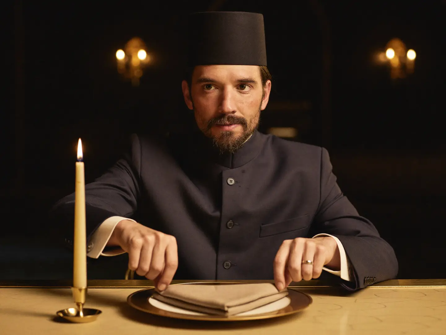 The maître d’hôtel setting a folded napkin on a gold-rimmed charger by candlelight