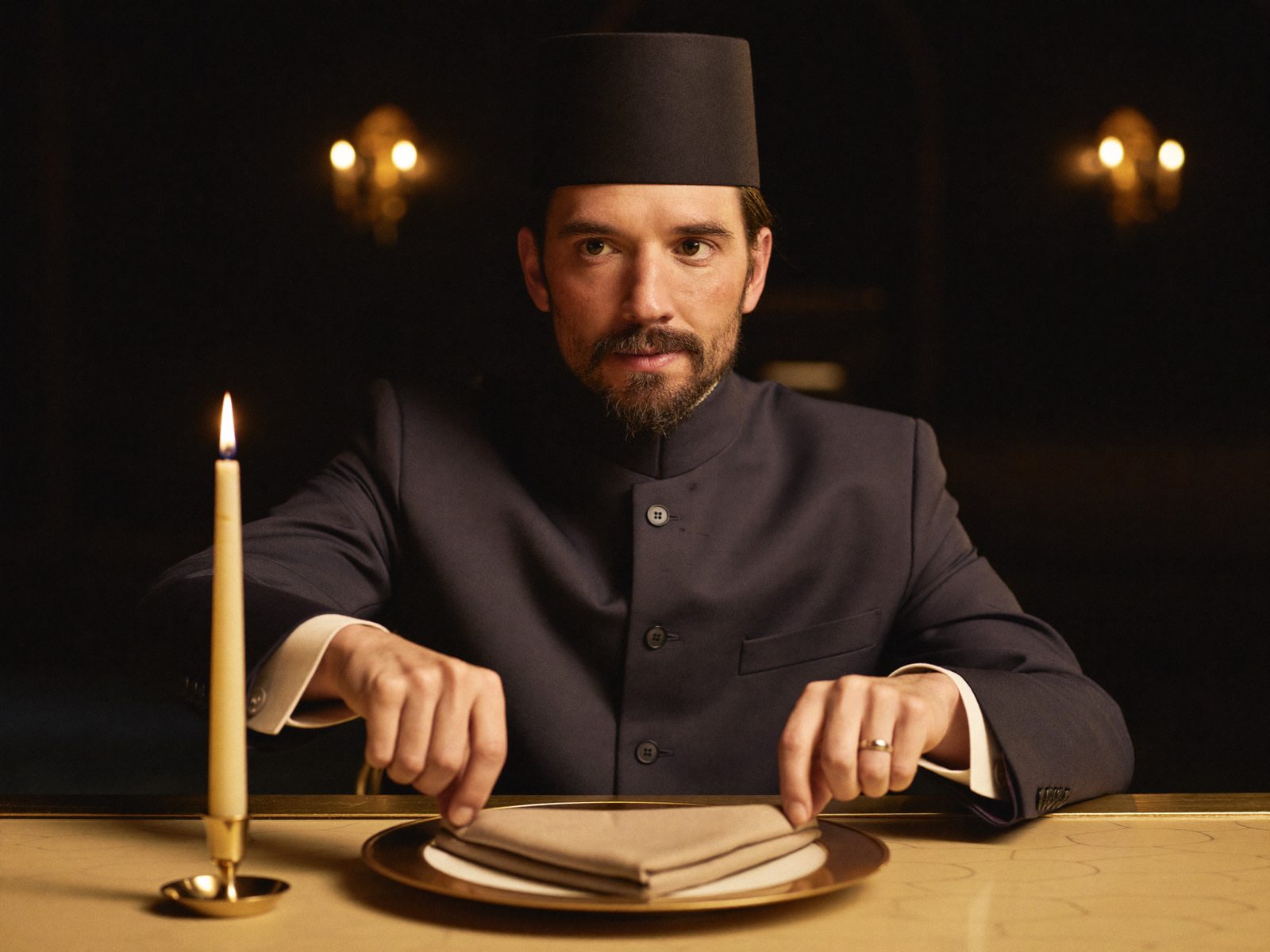 The maître d’hôtel setting a folded napkin on a gold-rimmed charger by candlelight