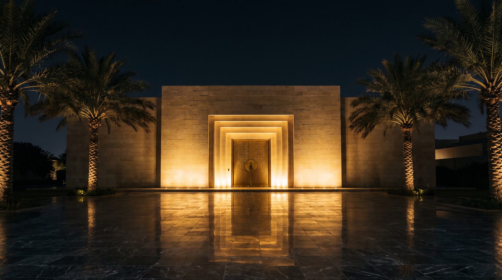 The hall at night — a limestone facade with a single lit brass portal and date palms in silhouette