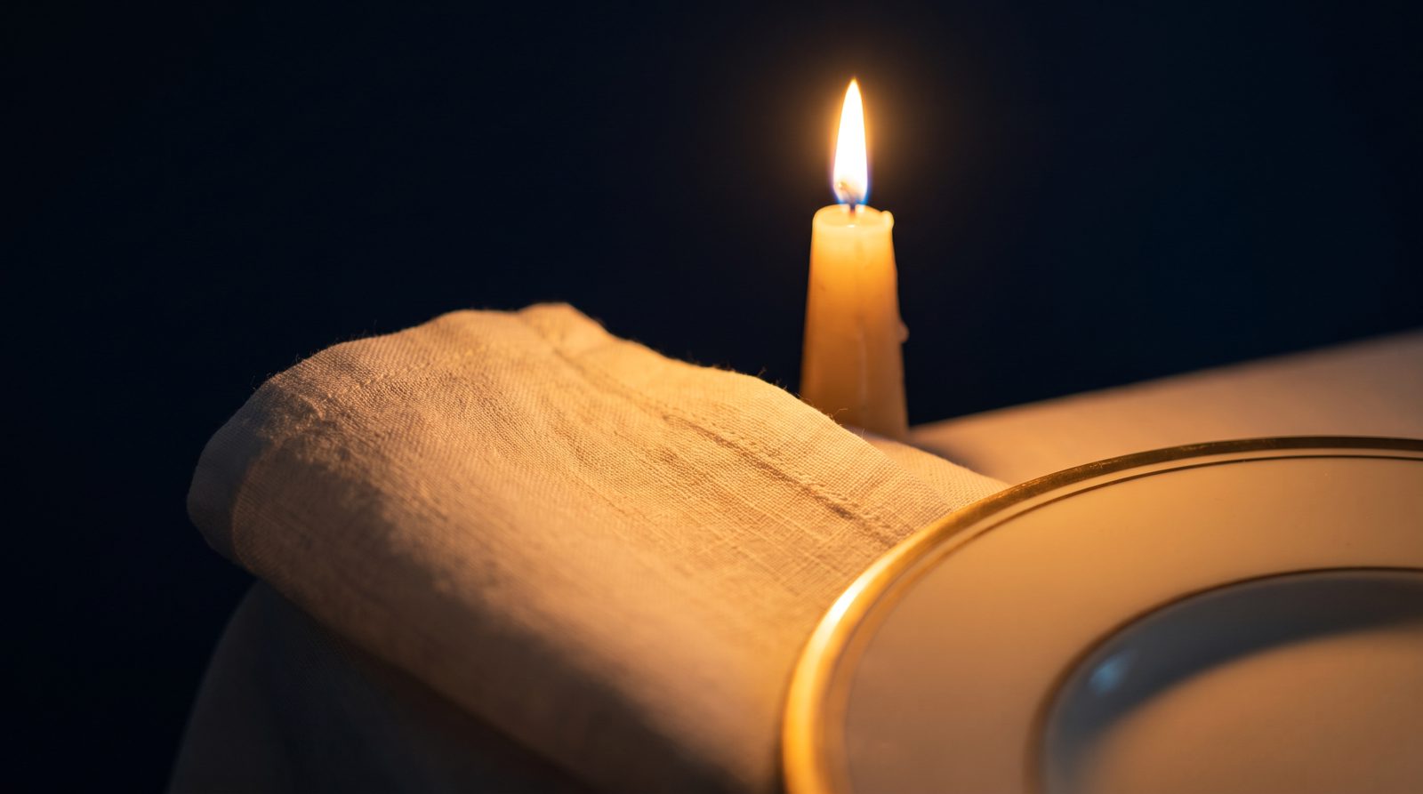 A gold-rimmed charger plate, folded linen and a tapered candle on ivory table linen